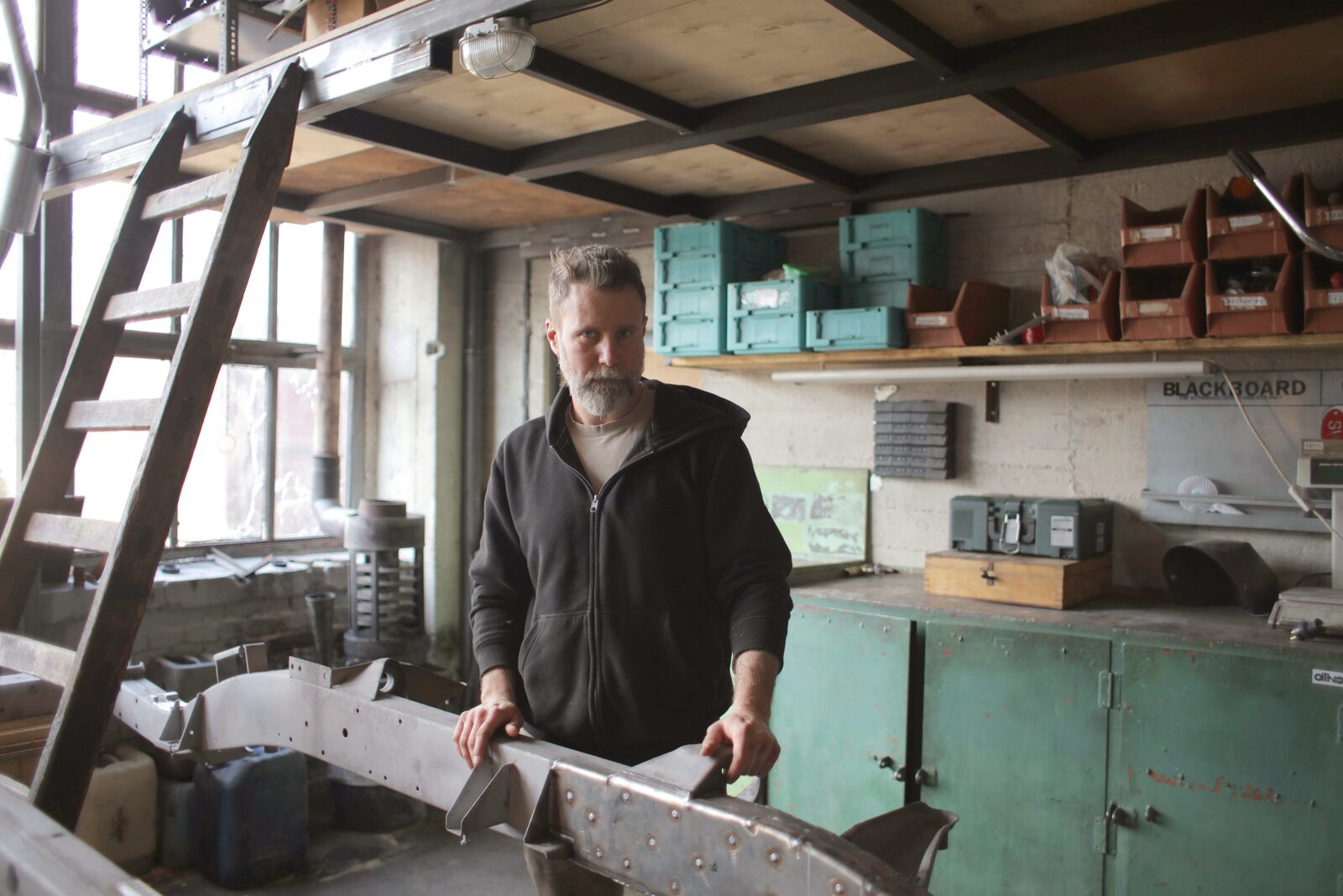 A business owner standing at his workbench in his workshop, beside a partly built metal frame, with tools and parts bins on the shelves behind him.